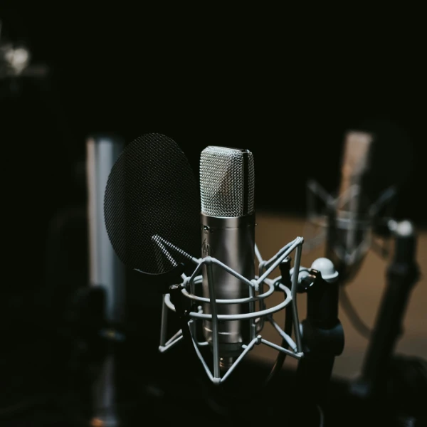 Close-up of a professional microphone in a recording studio. The microphone is mounted on a stand and equipped with a pop filter. The background is dark, and another microphone can be seen slightly blurred in the distance, creating a focused, intimate atmosphere typical of podcast.